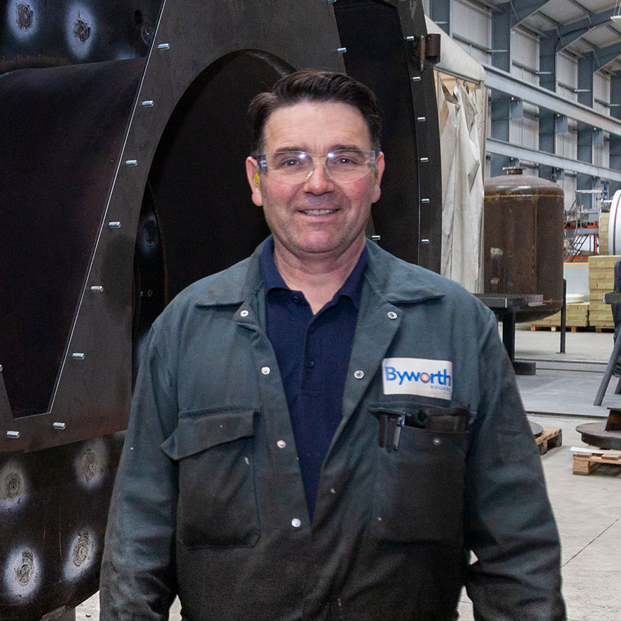 A man in a green boiler suit, smiling, he's in a factory. 
